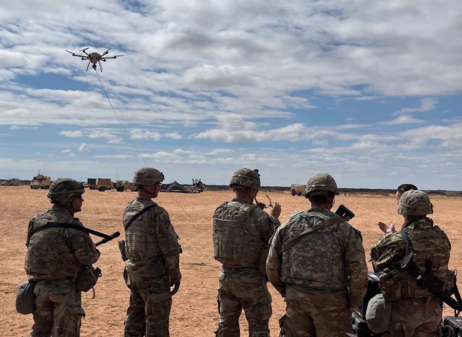 Soldiers in a desert area observe a tethered drone flying overhead, maintaining a vigilant watch.